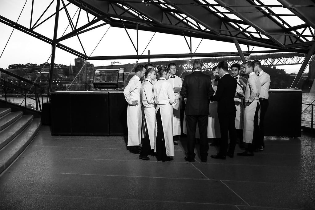 A group of waitstaff in white shirts and aprons stand in a circle around a man in a suit, having a discussion inside a modern glass building overlooking a bridge and water.