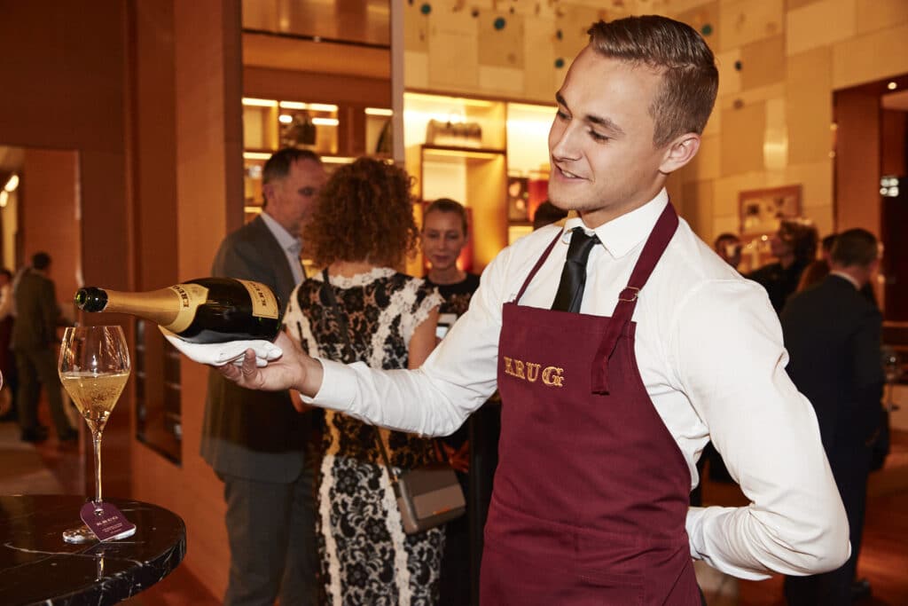A smiling server in a maroon apron pours champagne into a glass at a stylish indoor event, with elegantly dressed people socializing in the background.